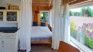 Ein Schlafzimmer mit Bett, einem Fenster mit Vorhängen, einer Holzbalkendecke und Blick auf Bäume und Blumen vor dem Fenster.