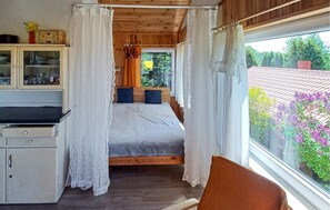 Ein Schlafzimmer mit Bett, einem Fenster mit Vorhängen, einer Holzbalkendecke und Blick auf Bäume und Blumen vor dem Fenster.