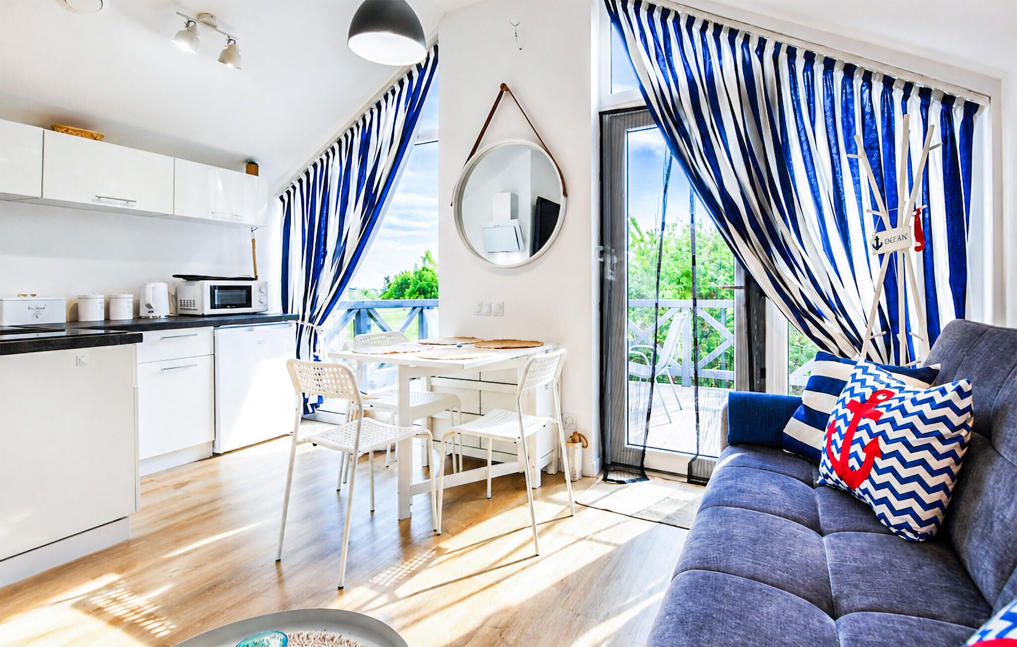A modern kitchen with white cabinets, a dining table with chairs, a gray sofa with patterned pillows, and blue and white striped curtains.