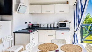 Una kitchenette compacta con microondas, hervidor y recipientes de almacenamiento. Hay sillas blancas y una mesa con individuales tejidos.