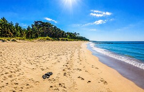 Ein Strand mit Fußabdrücken und einem Paar Sandalen, ein klarer Himmel und ein weiter in der Ferne liegender Steg.