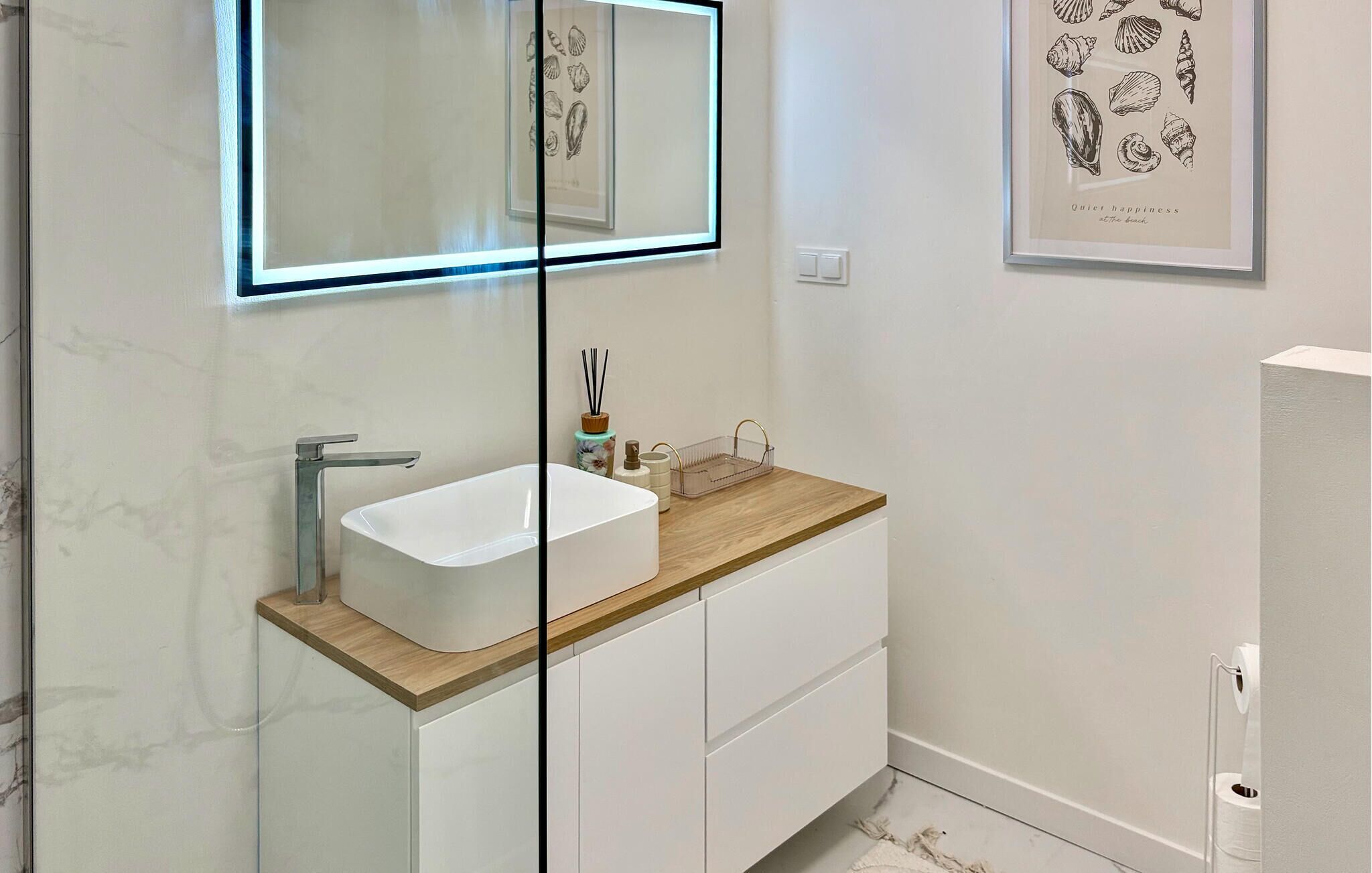 A modern bathroom with a white vanity, a glass-enclosed mirror, and a framed seashore illustration on the wall.