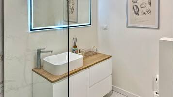 A modern bathroom with a white vanity, a glass-enclosed mirror, and a framed seashore illustration on the wall.
