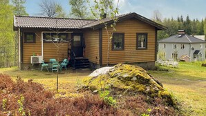 En stuga av trä med en veranda, utomhusmöbler och en stor sten täckt av mossa.