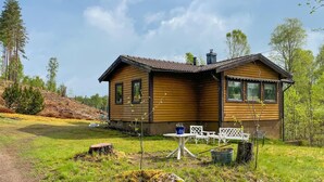 En stuga av trä med en skorsten, en liten uteplats med ett bord och stolar samt en gräsyta runt omkring.