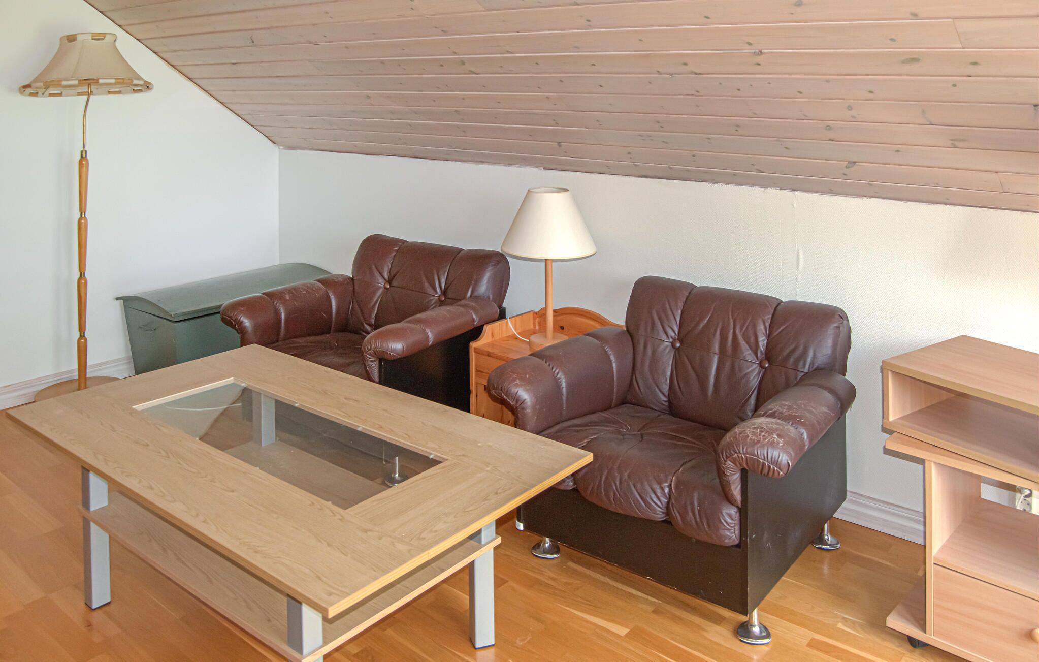 A room with a wooden ceiling, a glass-top table, two leather chairs, a lamp, and a wooden cabinet.