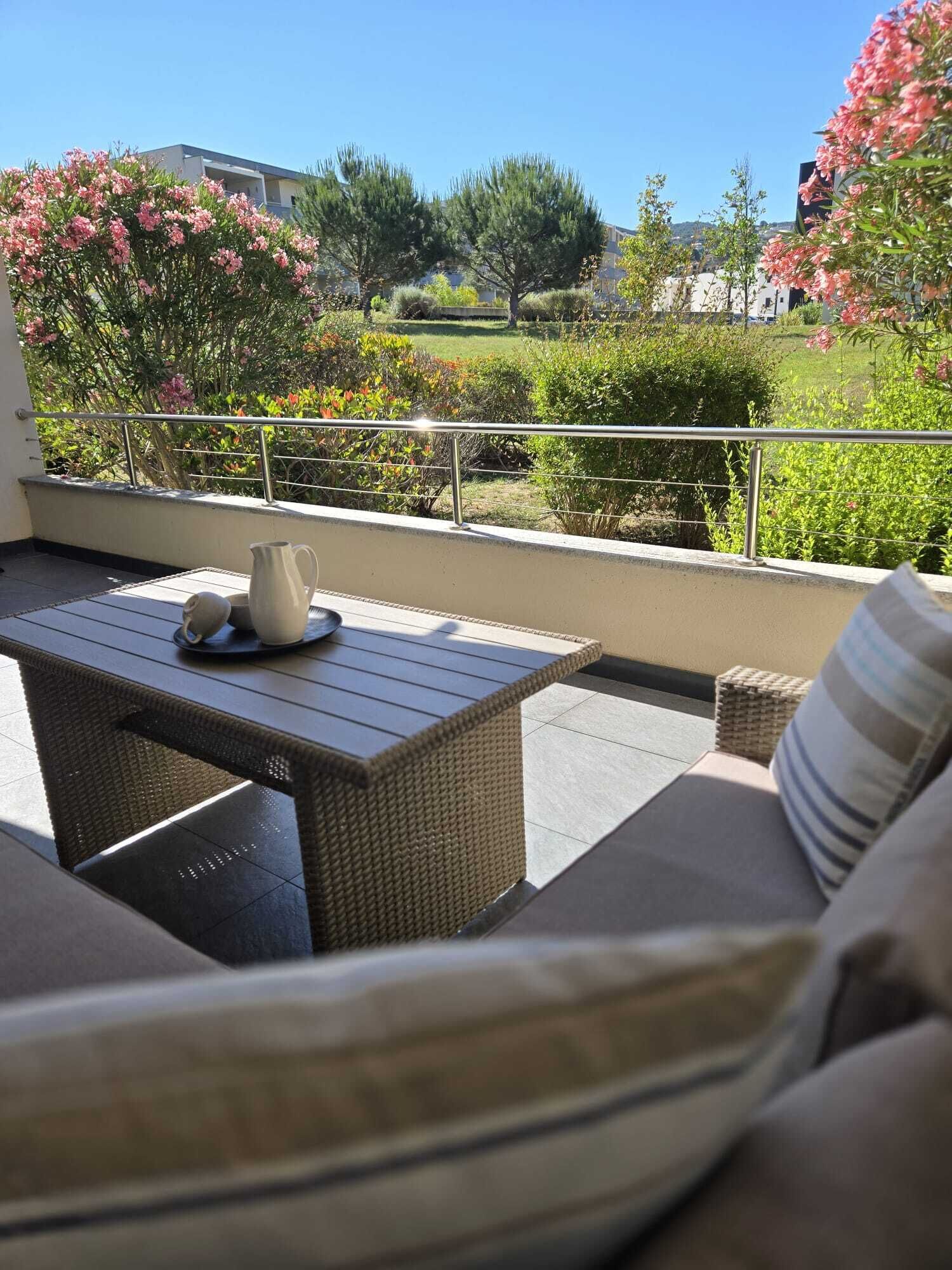 Ein Balkon im Freien mit Korbmöbeln, einem Metallgeländer und Blick auf einen Garten mit verschiedenen Pflanzen und Blumen.