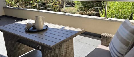 Uma varanda externa com móveis de vime, corrimão de metal e vista para um jardim com diversas plantas e flores.