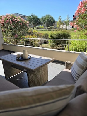 Ein Balkon im Freien mit Korbmöbeln, einem Metallgeländer und Blick auf einen Garten mit verschiedenen Pflanzen und Blumen.
