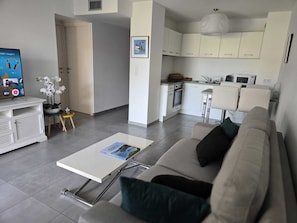 Ein modernes Wohnzimmer mit Sofa, Couchtisch und Fernseher. Es gibt eine Küchenzeile mit Schränken und einer Mikrowelle.
