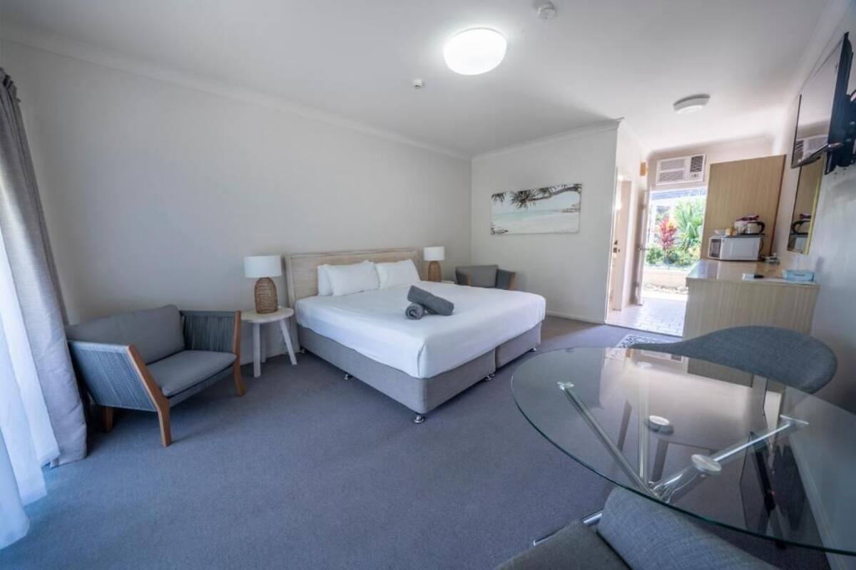 A hotel room with a bed, a glass table, a chair, a small table with a lamp, and a view of the outside.