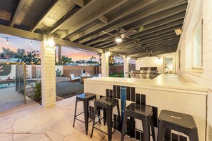 Ein Außenküchenbereich mit Barhocker-Bestuhlung, einem Deckenventilator und Blick auf den Garten.