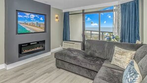 Una sala de estar con una televisión de pantalla plana, chimenea, sofá gris y un ventanal con vistas a la playa.
