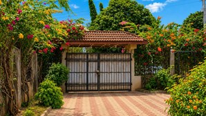 Una entrada porticada con techo de tejas, rodeada de abundante follaje y plantas con flores.