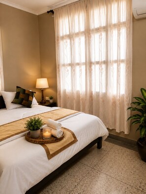 Habitación de hotel con dos camas, una mesita de noche con lámpara, una planta en maceta, una bandeja con vela y una ventana con cortinas translúcidas.