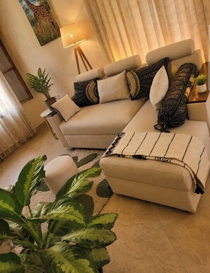 Una sala de estar con un sofá color beige, cojines estampados, una lámpara de pie y plantas verdes.