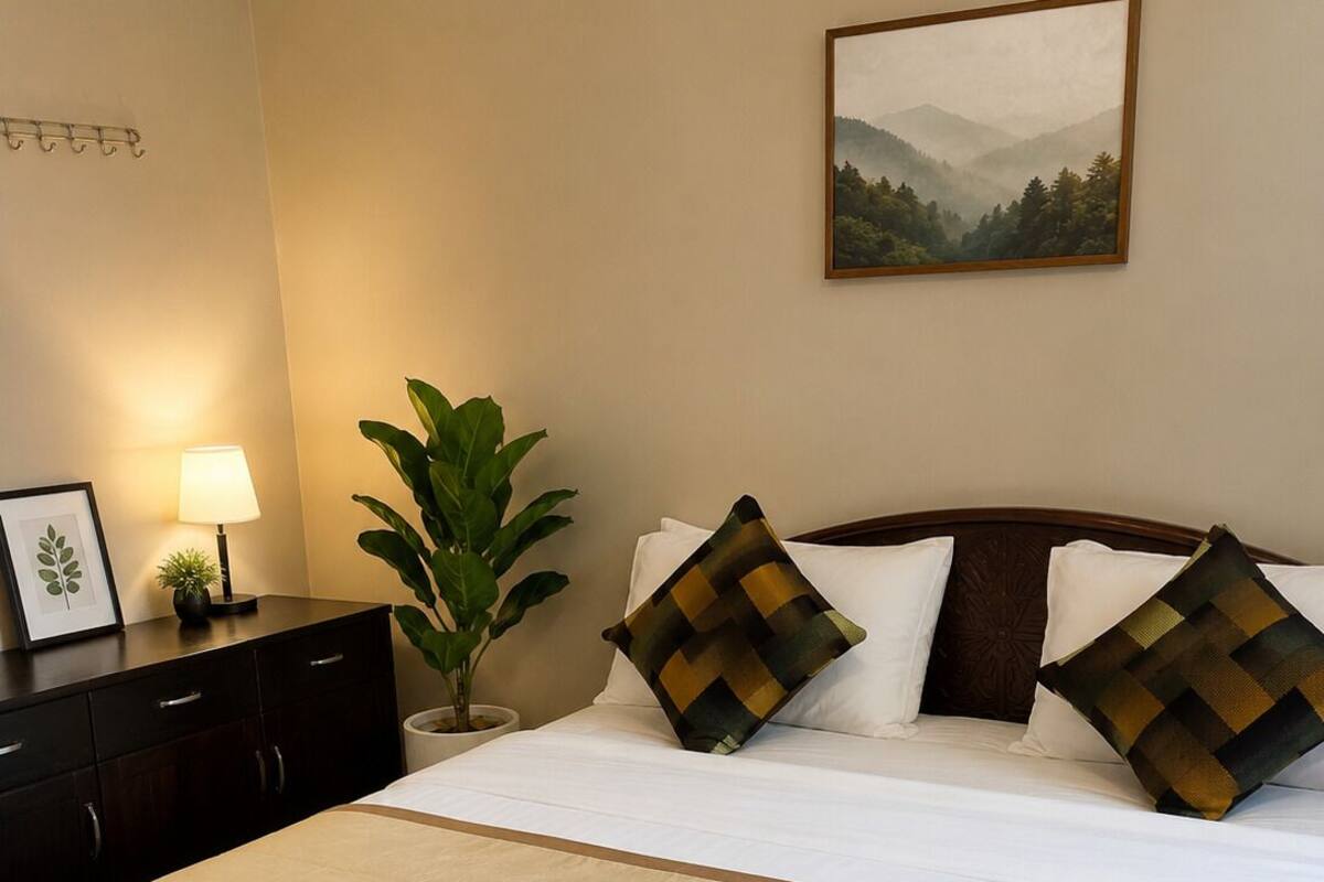 Una cama bien hecha con edredón blanco y dos almohadas decorativas, una mesita de noche con una lámpara, un cuadro enmarcado en la pared y una planta en maceta.