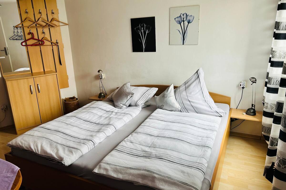 A double bed with striped bedding, a wooden wardrobe with hangers, a bedside table with a lamp, and two wall art pieces.