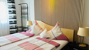 A hotel room with a bed, two bedside tables with lamps, a hanging coat rack, and a window with curtains. - Pension Merkle (Ichenhausen)