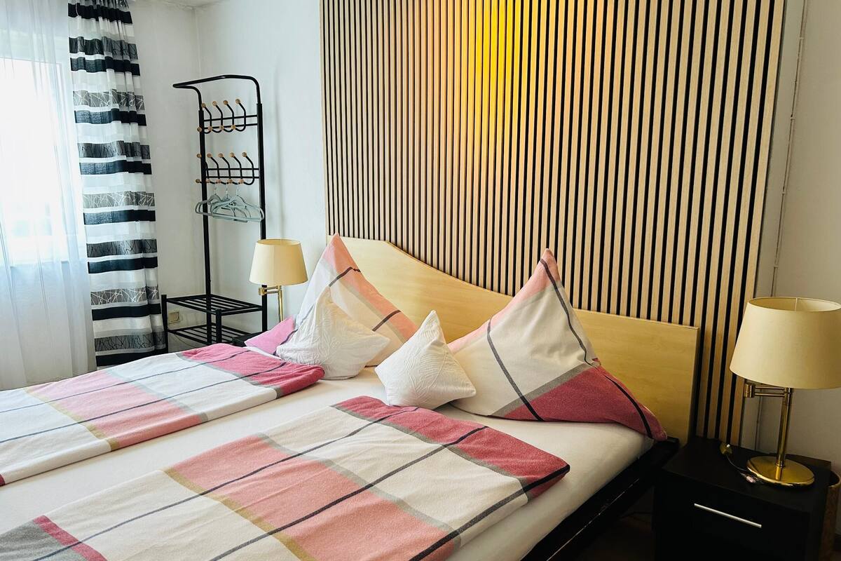 A hotel room with a bed, two bedside tables with lamps, a hanging coat rack, and a window with curtains.