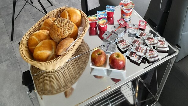 A dining area with a table set for breakfast, including bread, fruits, and packaged snacks. - Pension Merkle (Ichenhausen)