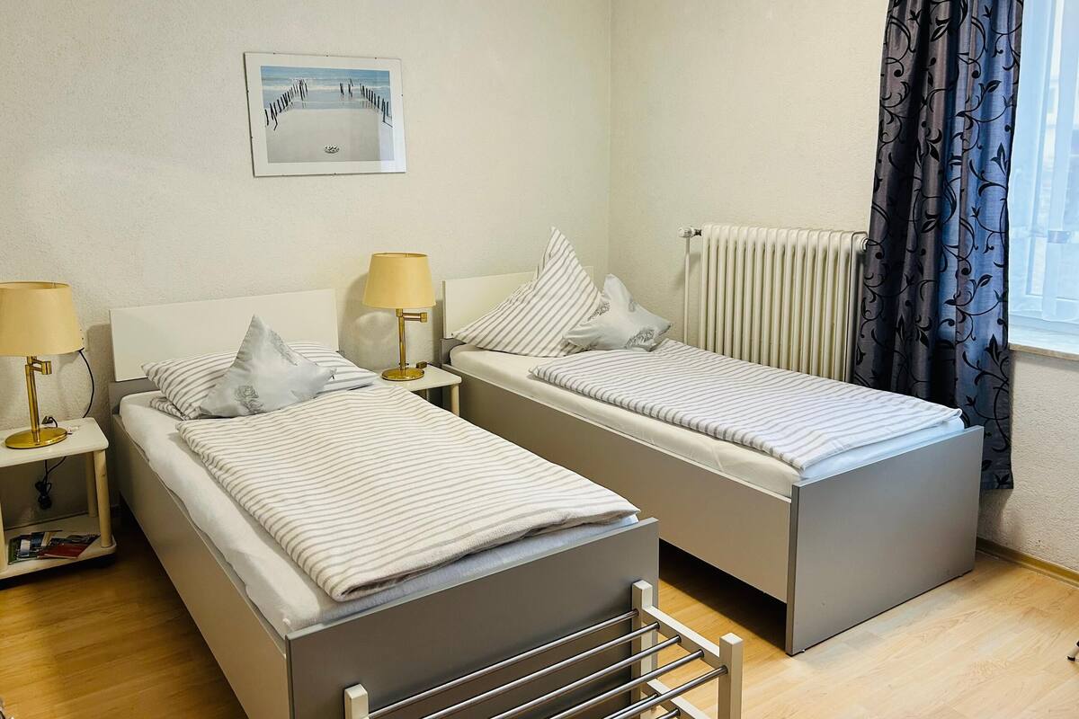 A twin room with two beds, bedside tables with lamps, a radiator, a framed picture on the wall, and a window with curtains.