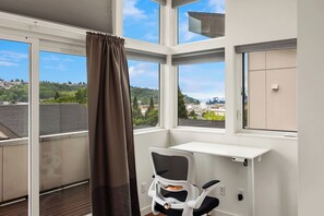 Een kamer met een groot raam, een stoel, een bureau en uitzicht op een stadsgezicht.