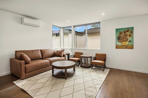 Een woonkamer met een bruine leren bank, twee houten stoelen, een ronde salontafel en een schilderij van zonnebloemen aan de muur.