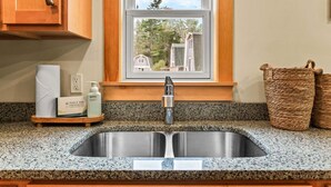 A kitchen with a stainless steel sink, wooden cabinets, a window with a view of houses, and a woven basket on the counter. - Four Lanterns - NEW! 4 Houses in Blue Hill (Blue Hill)