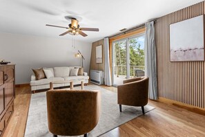 A modern living room with a ceiling fan, a sofa, two armchairs, a wooden cabinet, and a large window with a view of trees. - Four Lanterns - NEW! 4 Houses in Blue Hill (Blue Hill)