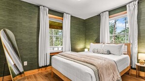 A bedroom with a large bed, two windows with curtains, a bedside table with a lamp, and a mirror on the wall. - Fern Cottage - NEW Cottage in Blue Hill! (Blue Hill)