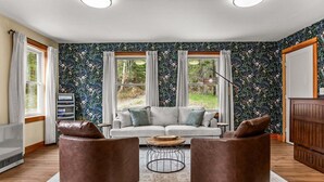 A living room with a sofa, two leather ottomans, a coffee table, and a large window with curtains. - Fern Cottage - NEW Cottage in Blue Hill! (Blue Hill)