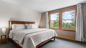 Ett sovrum med en sänggavel i trä, ett stort fönster med gardiner, en sänglampa och utsikt över träd.