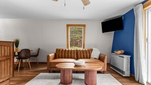 A modern living room with a leather sofa, a wooden coffee table, a TV mounted on the wall, and a large window with a view of trees. - Drift A-Frame - NEW A-Frame in Blue Hill! (Blue Hill)
