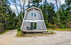 A small, single-story cabin with a porch and two chairs, surrounded by dense forest.