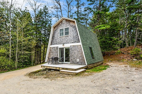 A small, single-story cabin with a sloped roof, a front porch with a table and chairs, and a door leading to the interior.. Drift A-Frame - NEW A-Frame in Blue Hill!