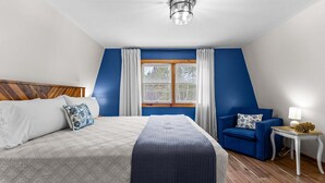 A bedroom with a bed, a blue armchair, a small table with a lamp, and a window with curtains. - Drift A-Frame - NEW A-Frame in Blue Hill! (Blue Hill)