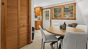 Un coin repas avec une table ronde, des chaises blanches, une fenêtre donnant sur des arbres et des meubles de rangement en bois.