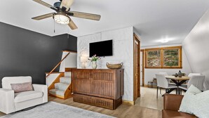 Un salon moderne avec un escalier en bois, un téléviseur à écran plat et un coin repas baigné de lumière naturelle.