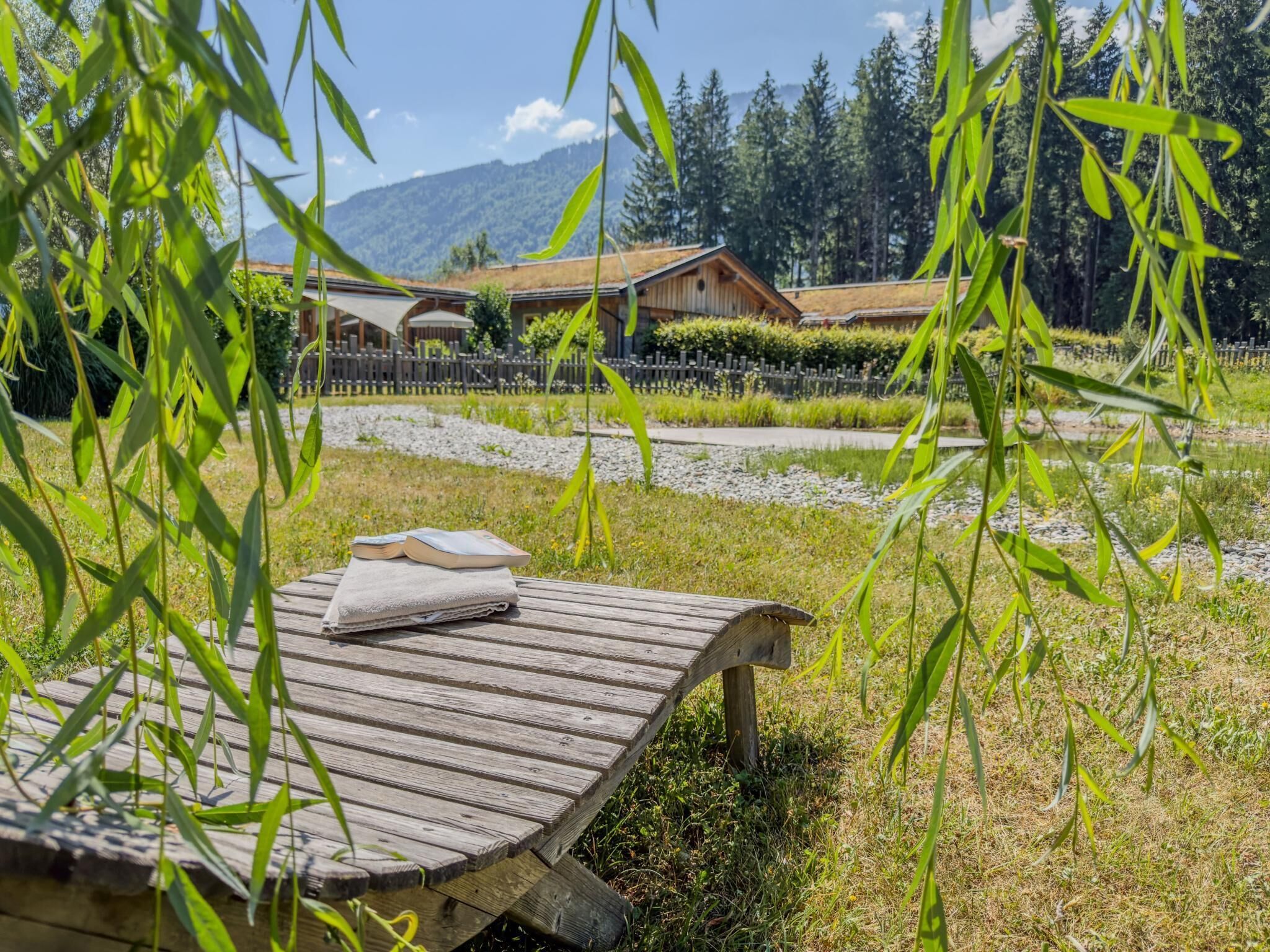 A wooden deck with a towel and book, surrounded by greenery and a mountain view.