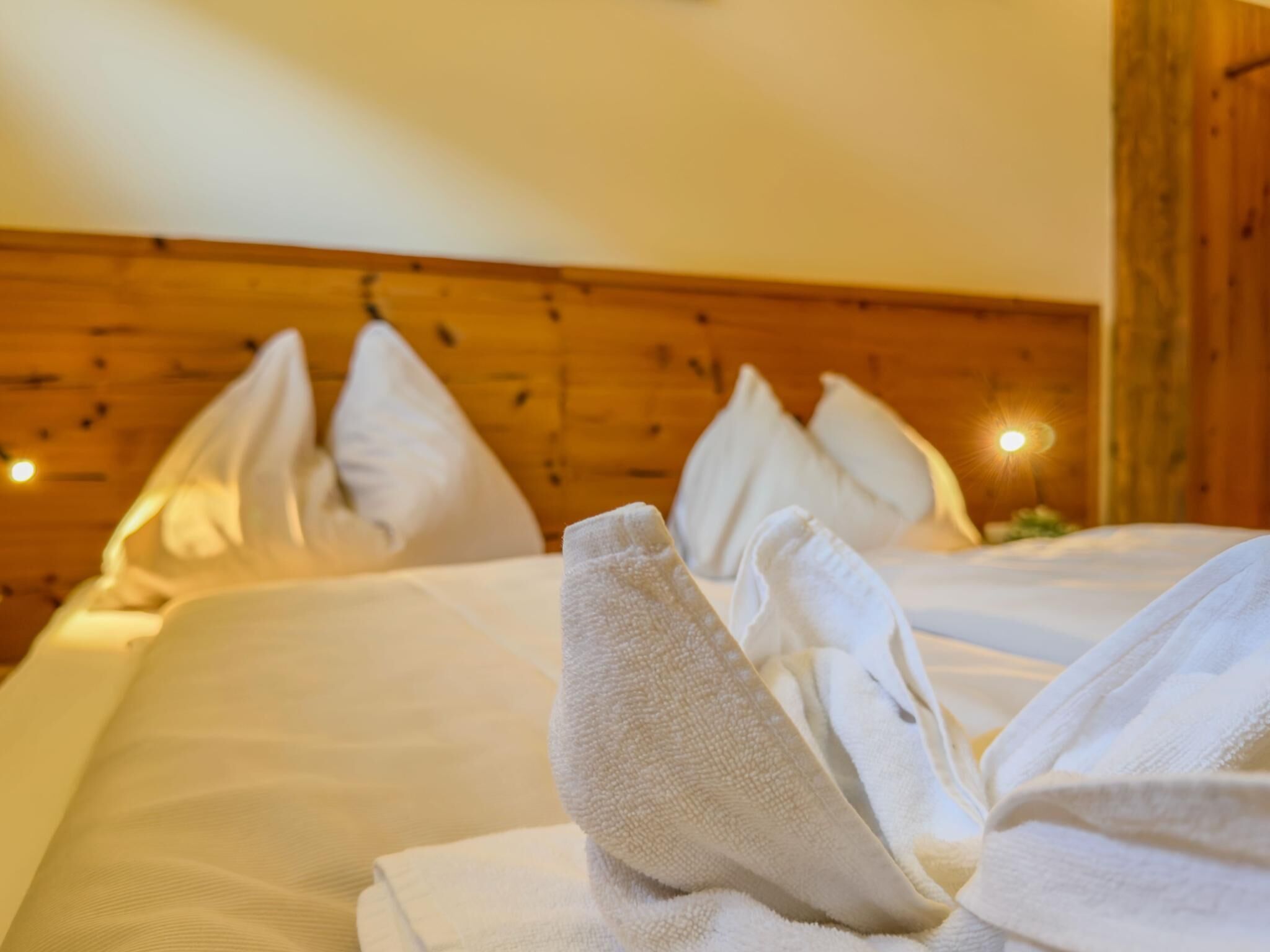 Cama bem arrumada com almofadas e toalhas dobradas, cabeceira de madeira e iluminação ambiente aconchegante.