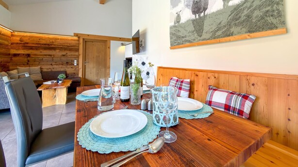 Un coin repas avec une table en bois dressée pour deux personnes, un banc avec des coussins à carreaux et un tableau représentant des moutons accroché au mur.