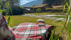 Une personne est allongée sur un lit, couverte d’une couverture à carreaux, porte un chapeau et tient un livre. Elle surplombe une zone herbeuse, délimitée par une clôture en bois, avec un bâtiment en bois et une montagne en arrière-plan.
