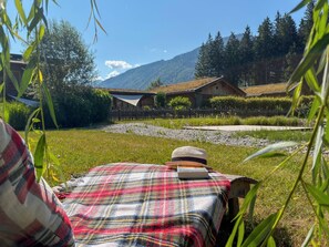 A person reclining on a bed with a plaid blanket, a hat, and a book, overlooking a grassy area with a wooden fence, a wooden building, and a mountain in the background. - Chalet with sauna near Nassfeld-Pressegger See (Hermagor-Pressegger See)