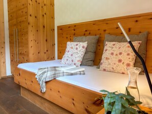 Ein hölzernes Bett mit einer weißen Matratze, mehreren Kissen mit floralem Muster, einer karierten Decke und einer klaren Glasvase mit einer Pflanze.