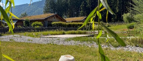 Ett träbord med en öppen bok och en halmhatt, placerat utomhus med utsikt över ett hus och en skog.