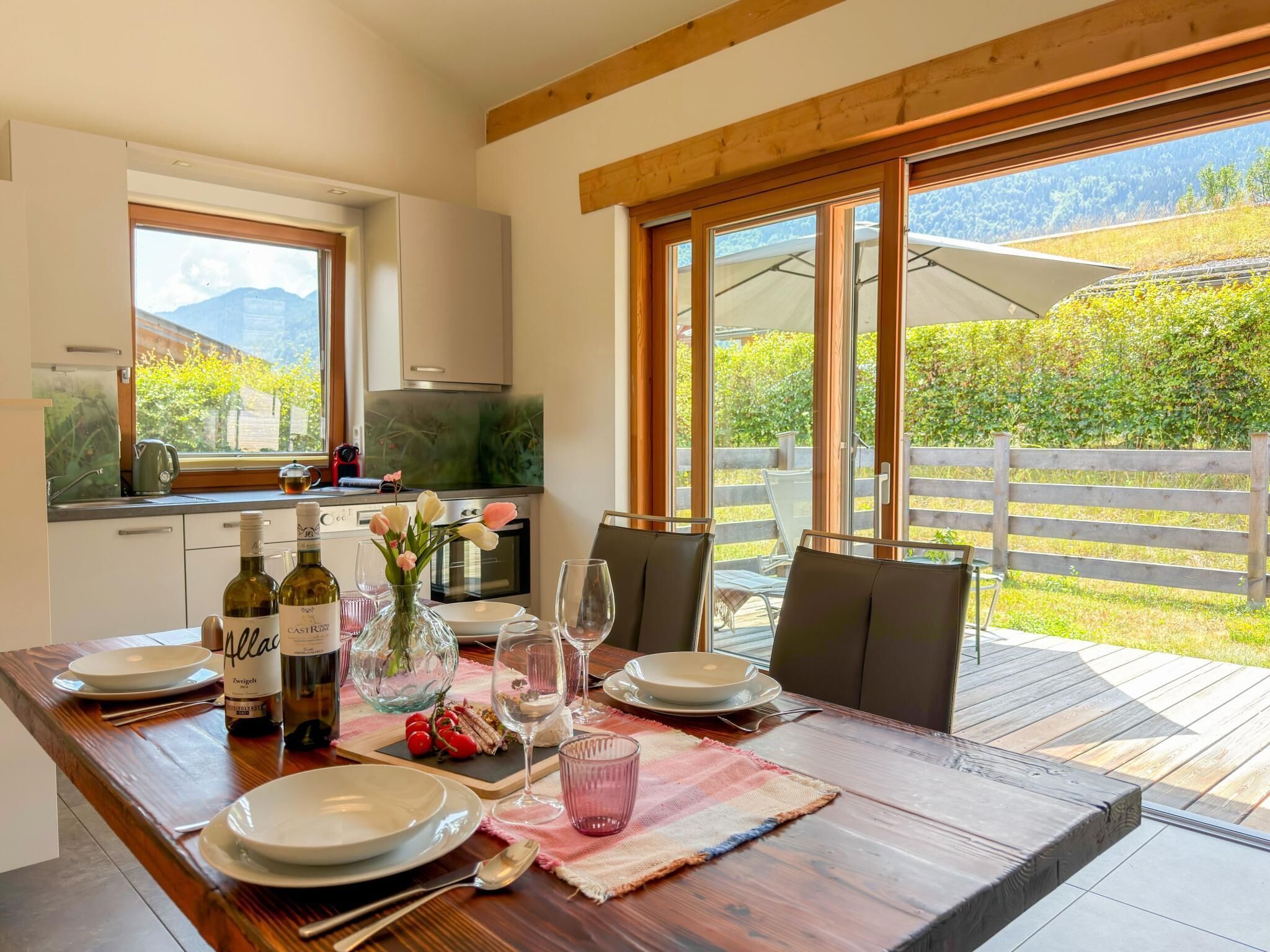 Un coin repas avec une table dressée pour deux personnes, une cuisine en arrière-plan et une vue sur un jardin par de grandes fenêtres.