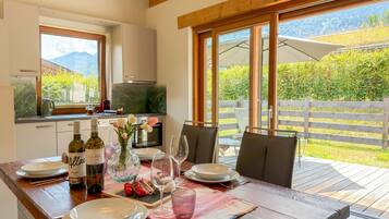 Un coin repas avec une table dressée pour deux personnes, une cuisine en arrière-plan et une vue sur un jardin par de grandes fenêtres.
