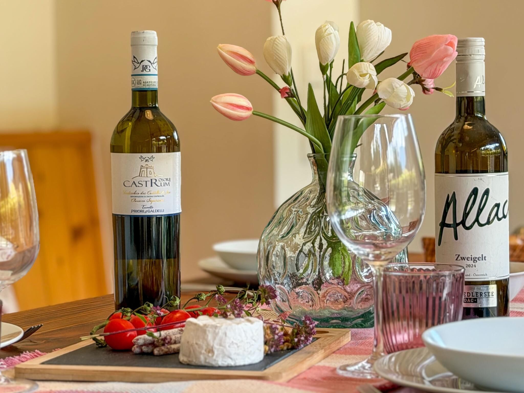 A table set with wine bottles, a vase of tulips, and a cheese board with tomatoes and flowers.