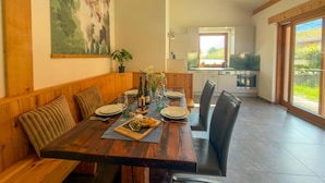Un comedor con una mesa de madera dispuesta para cuatro personas, un cuadro en la pared y una cocina al fondo.